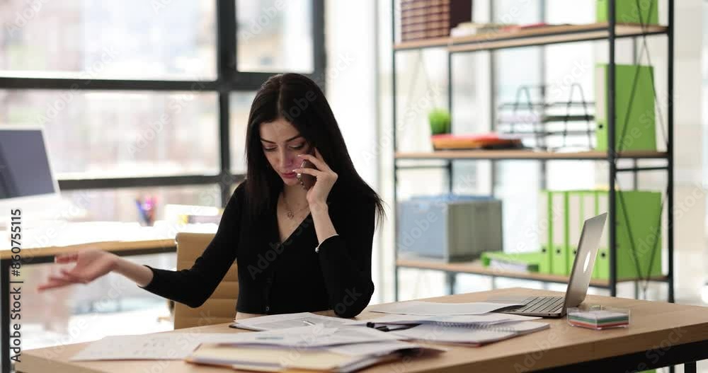 Woman talks on mobile phone with partner looking for document on wooden table. Elegant brunette manager at work in spacious modern office Woman talks on mobile phone with partner looking for document on wooden table. Elegant brunette manager at work in spacious modern office