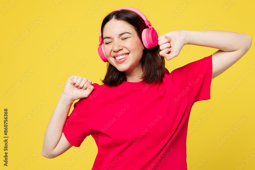 Young smiling happy cheerful woman wear pink t-shirt casual clothes listen to music in headphones raise up hands dance isolated on plain yellow orange background studio portrait. Lifestyle concept. Young smiling happy cheerful woman wear pink t-shirt casual clothes listen to music in headphones raise up hands dance isolated on plain yellow orange background studio portrait. Lifestyle concept.