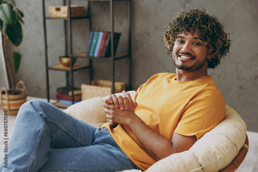 Φωτογραφία Side view young happy cool cheerful Indian man wear orange casual clothes look camera sits in grey chair stay at home hotel flat rest relax spend free spare time in living room indoor