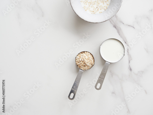 Top view of measuring cups with milk and oatmeal on a white marble table. Porridge of oat flakes.