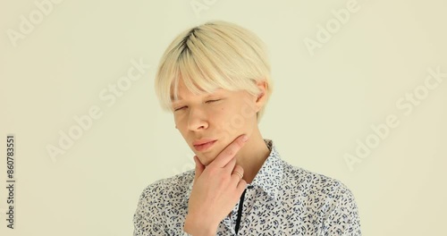 Blonde woman with short stylish hairdo suspiciously looks and squints. Lady in patterned blouse puts hand up to face while standing near wall photo