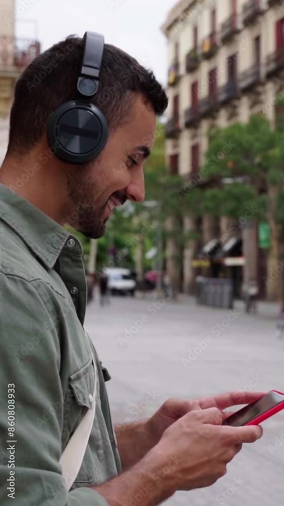 Cheerful caucasian man relaxing outdoors using phone sending text messages and checking the social media and listening music leaning in a brick wall in an urban street. Vertical footage. Cheerful caucasian man relaxing outdoors using phone sending text messages and checking the social media and listening music leaning in a brick wall in an urban street. Vertical footage.