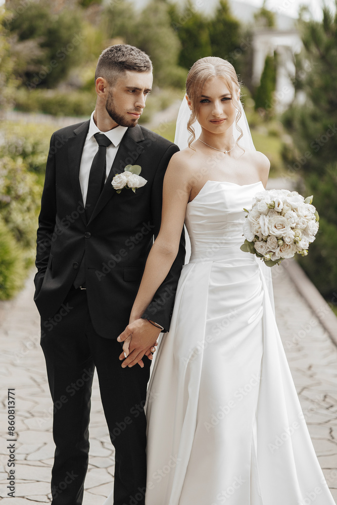 A bride and groom are standing together on a path, holding hands. The bride is wearing a white dress and the groom is wearing a black suit. Scene is happy and romantic
