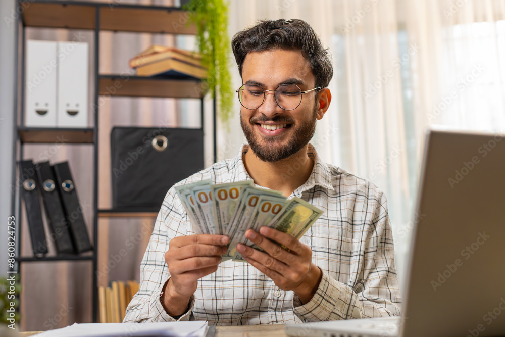Happy young Indian businessman counting money cash and using laptop in home office. Successful Arabian freelancer getting bundle of money in office, calculates dividends cash money profits finances. Happy young Indian businessman counting money cash and using laptop in home office. Successful Arabian freelancer getting bundle of money in office, calculates dividends cash money profits finances.