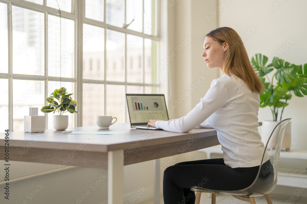 Young woman looking at notes while sitting at desk and typing on a computer showing a graph Young woman looking at notes while sitting at desk and typing on a computer showing a graph