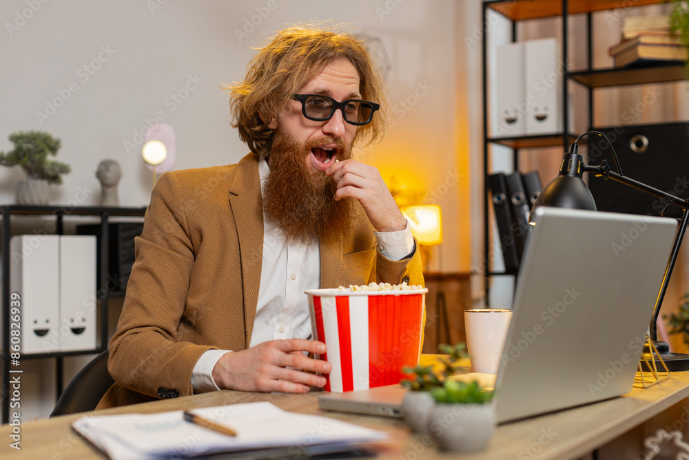 Caucasian businessman freelancer taking break from work wearing 3D glasses earing popcorn and watching movie on laptop. Smiling redhead man guy sitting at home office desk having snacks during break. Caucasian businessman freelancer taking break from work wearing 3D glasses earing popcorn and watching movie on laptop. Smiling redhead man guy sitting at home office desk having snacks during break.
