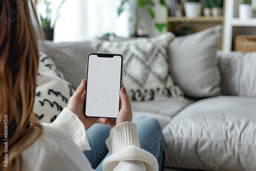 A woman is sitting on a couch holding a cell phone. The phone is blank, and the woman is looking at it. Concept of boredom or disinterest