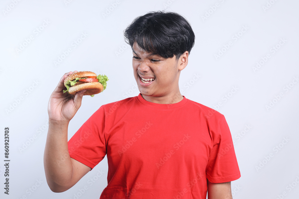 Potrait Of Annoyed Young Asian Male Raised Hand Holding And Looking Burger Isolated On White Background Potrait Of Annoyed Young Asian Male Raised Hand Holding And Looking Burger Isolated On White Background