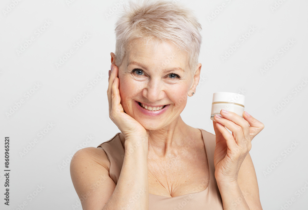 Smiling 60s middle aged mature woman putting tightening facial cream on face looking at camera. Smiling 60s middle aged mature woman putting tightening facial cream on face looking at camera.