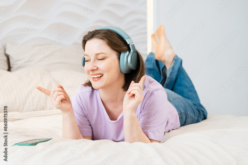 Woman lies on the bed in headphones and listens to music with her eyes closed and holding a smartphone in her hands Woman lies on the bed in headphones and listens to music with her eyes closed and holding a smartphone in her hands