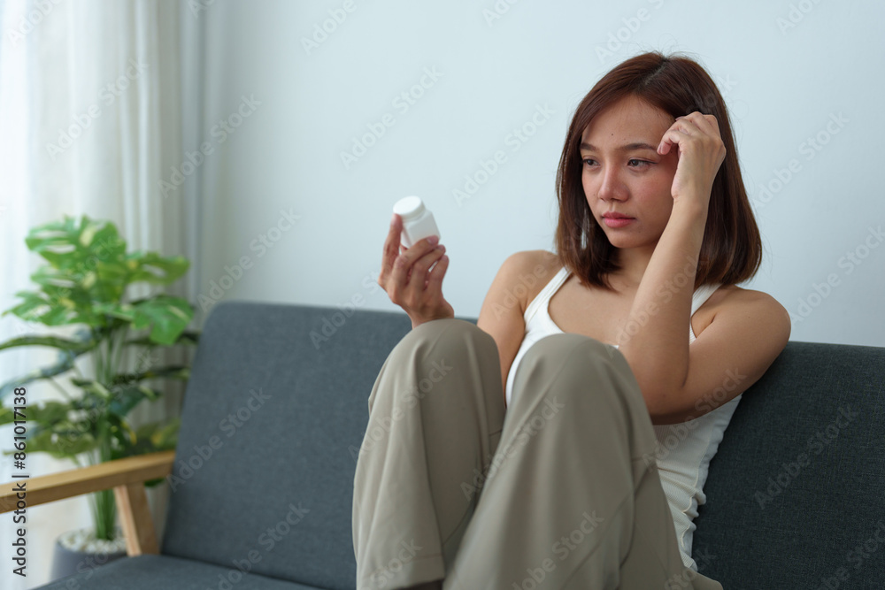 Young Asian woman suffering from work stress sits alone on the sofa at home, discouraged and takes a bottle of pills to relieve headache from fatigue. Desperate Asian woman feeling lonely. Young Asian woman suffering from work stress sits alone on the sofa at home, discouraged and takes a bottle of pills to relieve headache from fatigue. Desperate Asian woman feeling lonely.