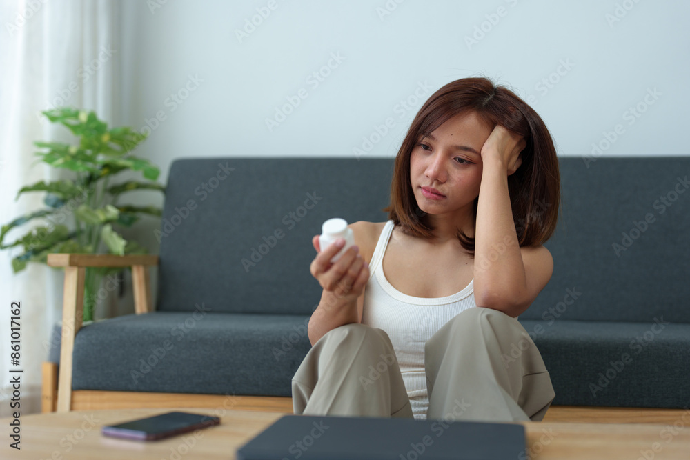 Young Asian woman suffering from work stress sits alone on the sofa at home, discouraged and takes a bottle of pills to relieve headache from fatigue. Desperate Asian woman feeling lonely.