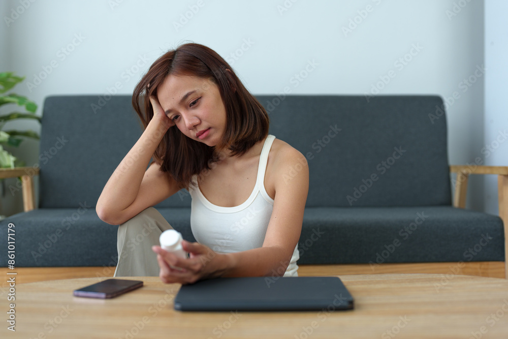 Young Asian woman suffering from work stress sits alone on the sofa at home, discouraged and takes a bottle of pills to relieve headache from fatigue. Desperate Asian woman feeling lonely. Young Asian woman suffering from work stress sits alone on the sofa at home, discouraged and takes a bottle of pills to relieve headache from fatigue. Desperate Asian woman feeling lonely.