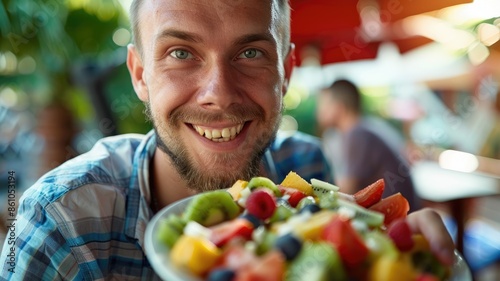 A bearded man is joyfully smiling, showcasing a bowl of fresh fruit salad at a vibrant outdoor market AIG58 photo