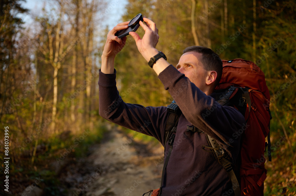 Fully equipped hiker taking pictures of sunrise on smartphone early in the morning in the mountains. Man standing with cell phone on mountain path enjoying sunrise. Fully equipped hiker taking pictures of sunrise on smartphone early in the morning in the mountains. Man standing with cell phone on mountain path enjoying sunrise.