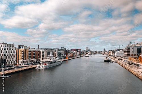 Urban Waterfront in Dublin with Luxury Yacht, Modern Architecture, Scenic River View, and Iconic Bridge under Partly Cloudy Sky