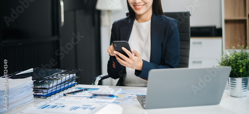 Young Asian woman, smiling and working at her desk in an office environment. She is wearing a business suit and holding documents