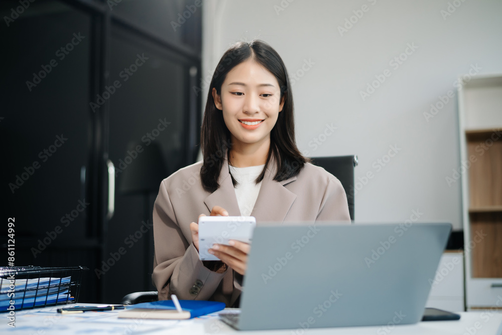 Asian woman, smiling and working at her desk in an office environment. She is wearing a business suit and holding documents while using a laptop. Asian woman, smiling and working at her desk in an office environment. She is wearing a business suit and holding documents while using a laptop.