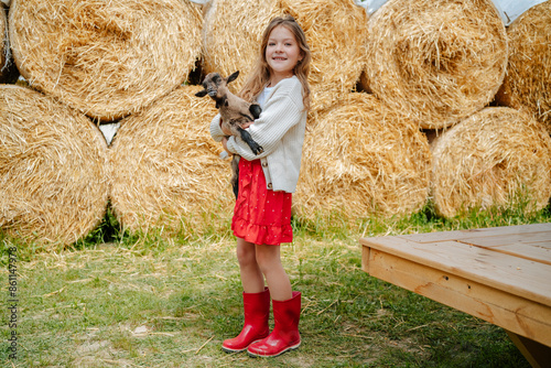 Cute girl carrying young goat standing in front of haystack at farm