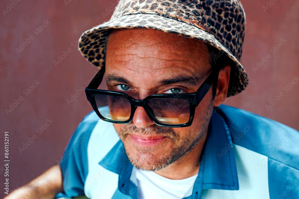 Smiling man wearing sunglasses and leopard print hat in front of wall Smiling man wearing sunglasses and leopard print hat in front of wall