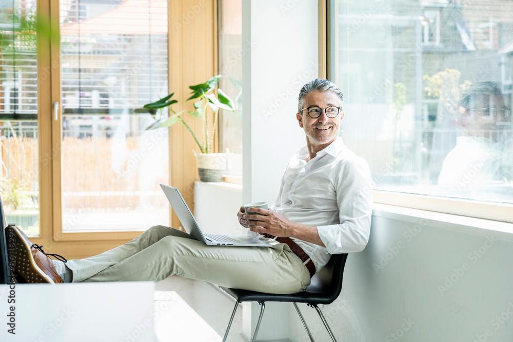 © Westend61 - Smiling businessman holding coffee cup using laptop working at office