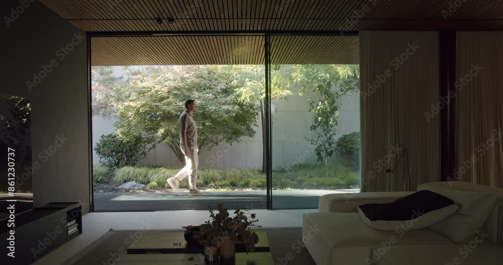 A peaceful image capturing a man strolling through a tranquil garden next to a modern living room. The natural light and lush greenery highlight quiet moments of solitude.