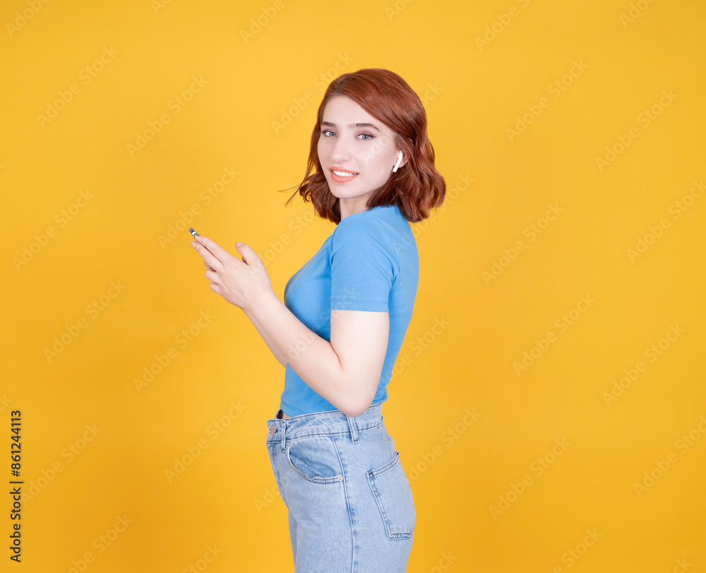 Obraz premium Side view portrait of young caucasian red bob hair 20s woman using mobile phone looking camera wear wireless earphones smiling satisfied. Using application, typing message chatting online.