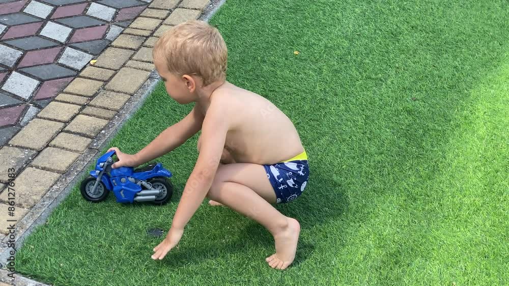 child in swimming trunks playing with toy motorcycle on artificial grass in courtyard of summer resort hotel