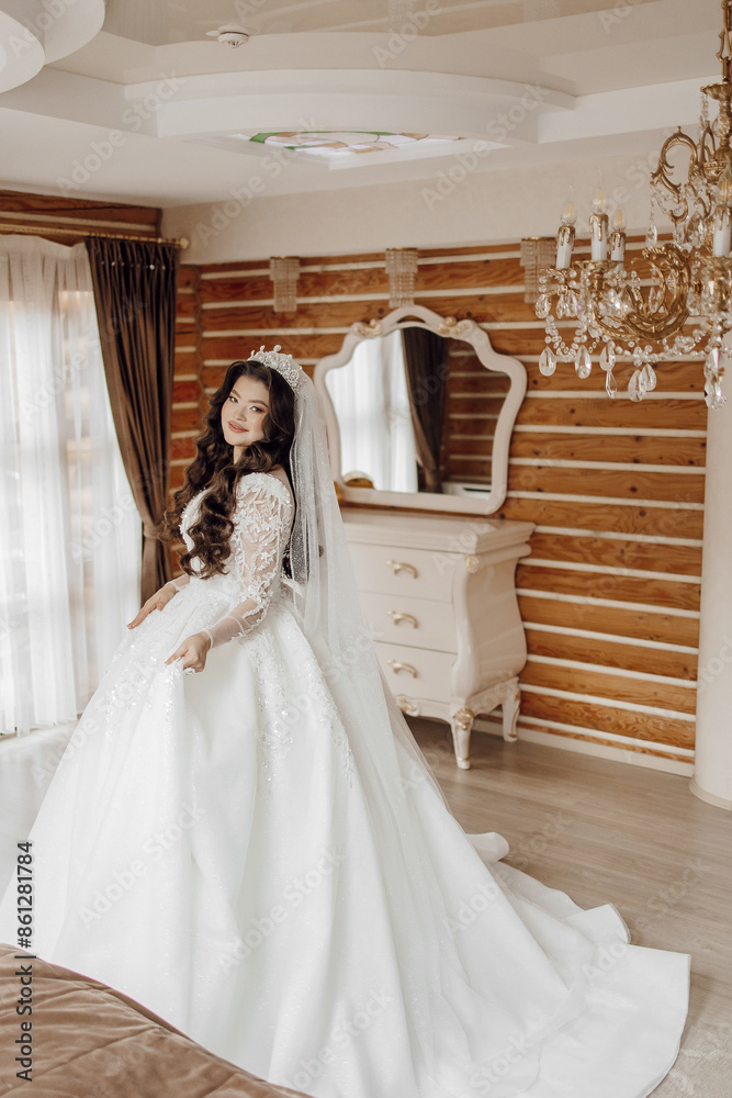 Fototapeta premium A woman in a long white dress stands in front of a mirror. She is wearing a tiara and a veil. The room is decorated with wooden furniture and a chandelier. Scene is elegant and sophisticated