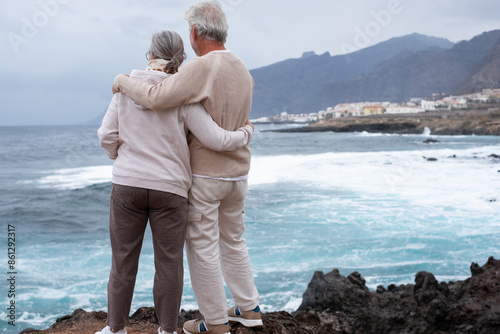 Rear view of a romantic senior couple embrace on the seashore on a bad weather day looking waves splashing against the coast enjoying retirement lifestyle or vacation © luciano