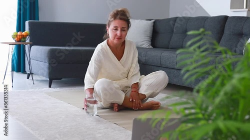 A beautiful middle-aged woman in a white outfit is drinking water afte yoga exersices photo