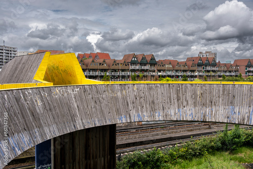 A scenic image showing Luchtsingel a striking wooden bridge with bright yellow accents, overlooking rustic houses with triangular roofs, under a cloudy sky, providing a fine balance of urban and natu