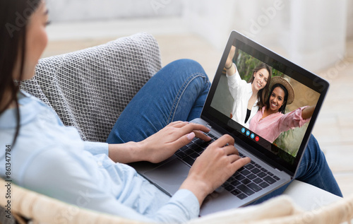 A woman is sitting on a couch at home, using a laptop to video chat with two friends. The friends are smiling and waving at the camera, while the woman is typing on the keyboard.