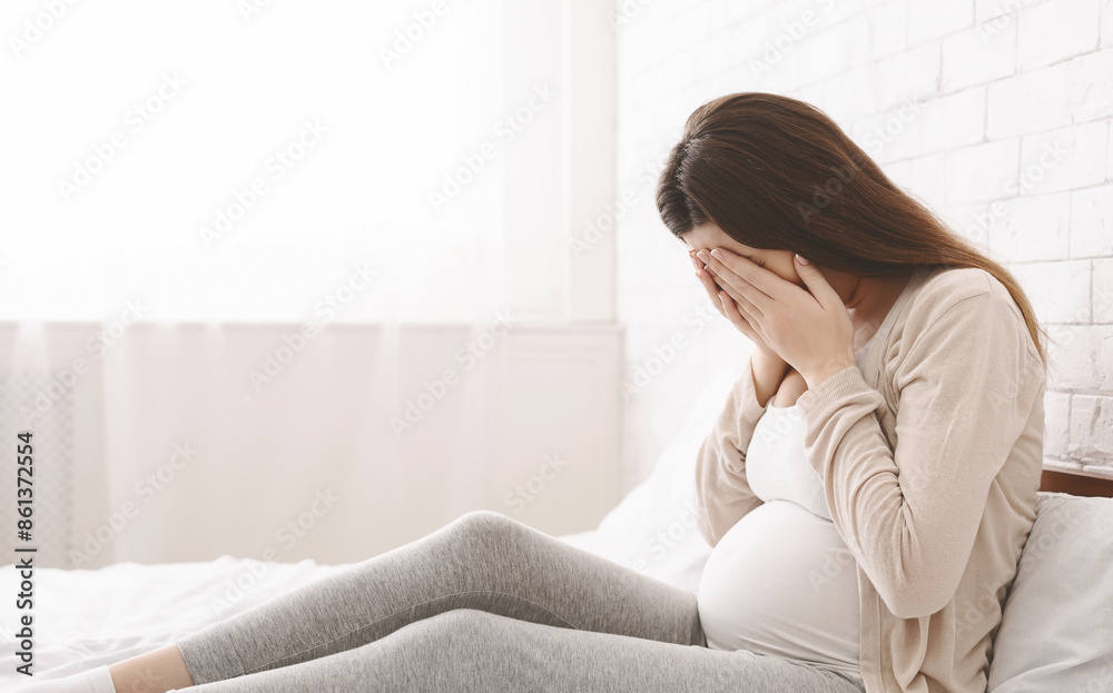 A pregnant woman sitting on a bed with her hands covering her face. She appears to be in a moment of contemplation or distress. A pregnant woman sitting on a bed with her hands covering her face. She appears to be in a moment of contemplation or distress.