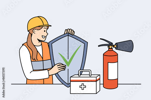 Woman safety specialist, stands near table with first aid kit and fire extinguisher and holds shield in hands. Girl safety inspector calls for caution, working to preserve life and health.