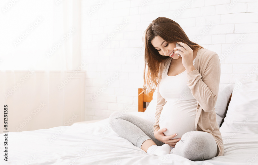 A pregnant woman is sitting on a bed, holding a cell phone to her ear while engaged in a phone conversation. She appears relaxed and comfortable as she talks on the phone in a cozy bedroom setting. A pregnant woman is sitting on a bed, holding a cell phone to her ear while engaged in a phone conversation. She appears relaxed and comfortable as she talks on the phone in a cozy bedroom setting.