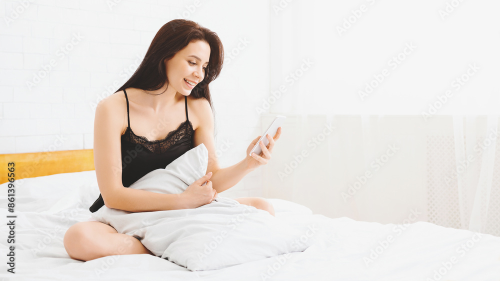 A woman is seated on a bed, holding a smartphone in her hands and looking at the screen. She appears focused on the device while sitting in a bedroom, copy space A woman is seated on a bed, holding a smartphone in her hands and looking at the screen. She appears focused on the device while sitting in a bedroom, copy space