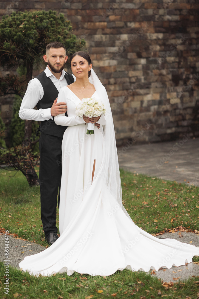 A bride and groom are posing for a picture in front of a brick wall. The bride is holding a bouquet of flowers and the groom is wearing a vest. Scene is happy and celebratory A bride and groom are posing for a picture in front of a brick wall. The bride is holding a bouquet of flowers and the groom is wearing a vest. Scene is happy and celebratory