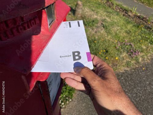 A hand puts an envelope for the United Kingdom General Election postal vote in a red post box.First Class.Envelope B