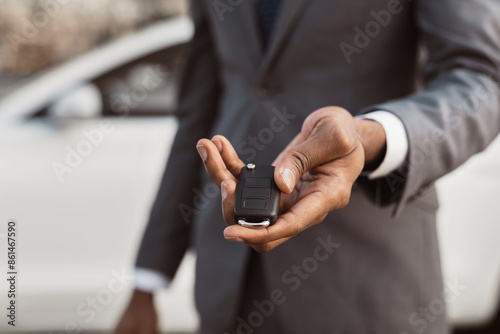 African American man wearing a gray suit holds a black car key in his right hand, palm up. He is standing in front of a white sedan, and the cars front side is out of focus. © Prostock-studio