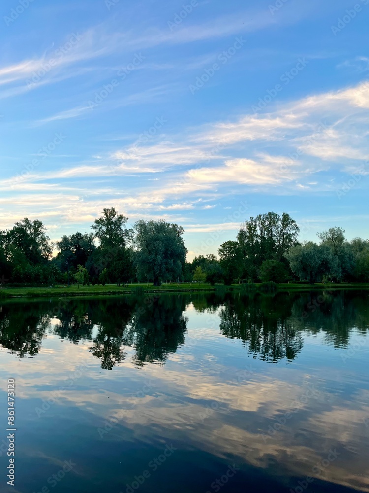 Sky and trees reflection on the calm surface of the lake, summer lake in the park, green trees, blue sky with tender clouds Sky and trees reflection on the calm surface of the lake, summer lake in the park, green trees, blue sky with tender clouds