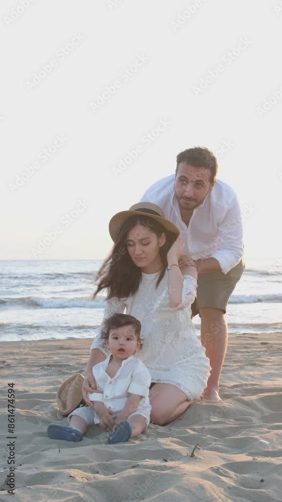 A baby boy in a white shirt and hat sits on the sand with his mother in a white dress behind him and his father smiling behind her. They enjoy a beautiful sunset by the sea A baby boy in a white shirt and hat sits on the sand with his mother in a white dress behind him and his father smiling behind her. They enjoy a beautiful sunset by the sea