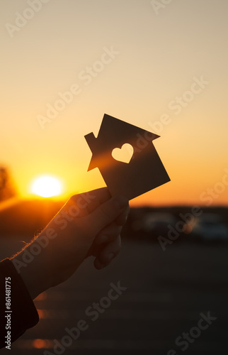 Silhouette of man's hand holding small wooden house against sunset.