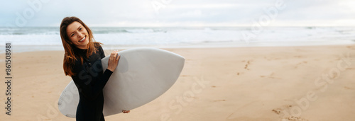 Sticker A woman in a black wetsuit stands on a sandy beach, holding a white surfboard under her arm