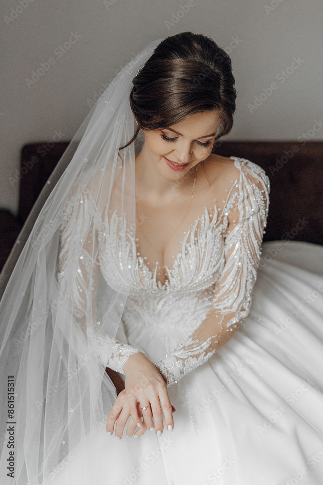 A woman is sitting on a bed wearing a white wedding dress. She is wearing a white veil and has her hands on her lap A woman is sitting on a bed wearing a white wedding dress. She is wearing a white veil and has her hands on her lap