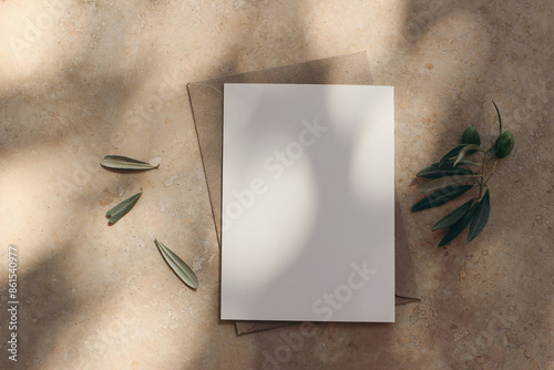 Mediterranean wedding stationerymockup in sunlight. Blank greeting card, envelope on beige marble stone floor background. Olive tree branch, fruit. Dappled light. Summer holiday design. Flatlay, top photo