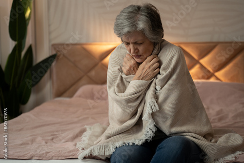 Fotografía A woman in her late 60s or early 70s is sitting on a bed, wrapped in a blanket, and looking unwell