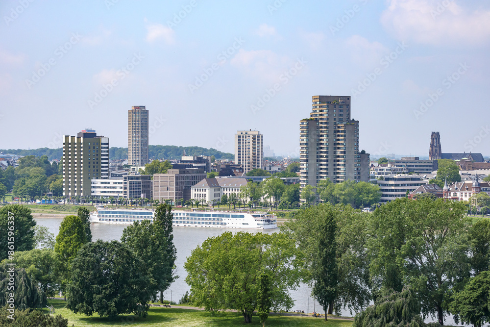 Rhine river with a tourist boat seen from high perspective, green leafed trees of Rheinpark public park in foreground, cityscape with buildings in background, cloudy spring day in Cologne, Germany Rhine river with a tourist boat seen from high perspective, green leafed trees of Rheinpark public park in foreground, cityscape with buildings in background, cloudy spring day in Cologne, Germany