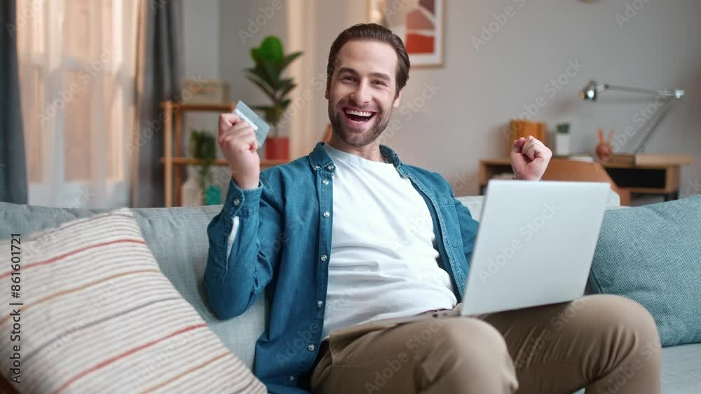 Cheerful man sitting on sofa and using laptop. Male sitting on couch and shopping online. Happy man successfully finishing internet purchase. Male holding credit card and rejoicing. Cheerful man sitting on sofa and using laptop. Male sitting on couch and shopping online. Happy man successfully finishing internet purchase. Male holding credit card and rejoicing.