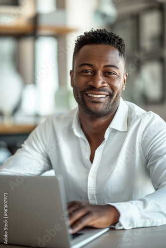 Attractive African American businessman sitting at his desk with a laptop, smiling and looking at the camera, wearing a white shirt, with short black hair and beard, against a light background.